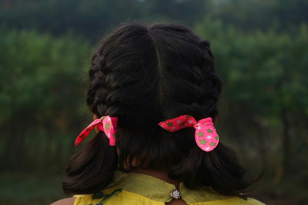 A view from behind of a girl with braided hair tied with pink star-patterned ribbons in a natural setting.