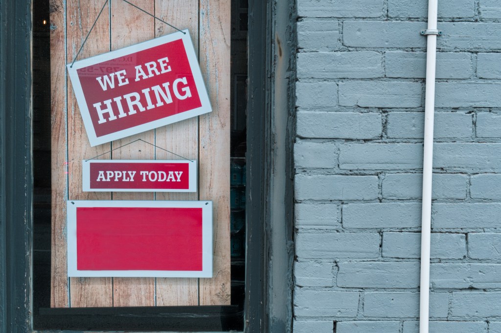 A storefront window displaying a 'We Are Hiring' sign in red and white, along with a smaller sign that says 'Apply Today.'