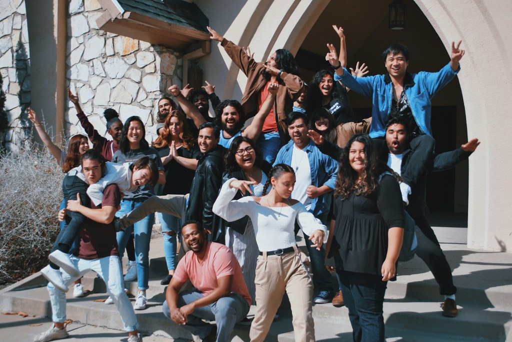 A large group of diverse individuals posing joyfully outdoors, some are jumping or making playful gestures, in front of a stone and stucco building.