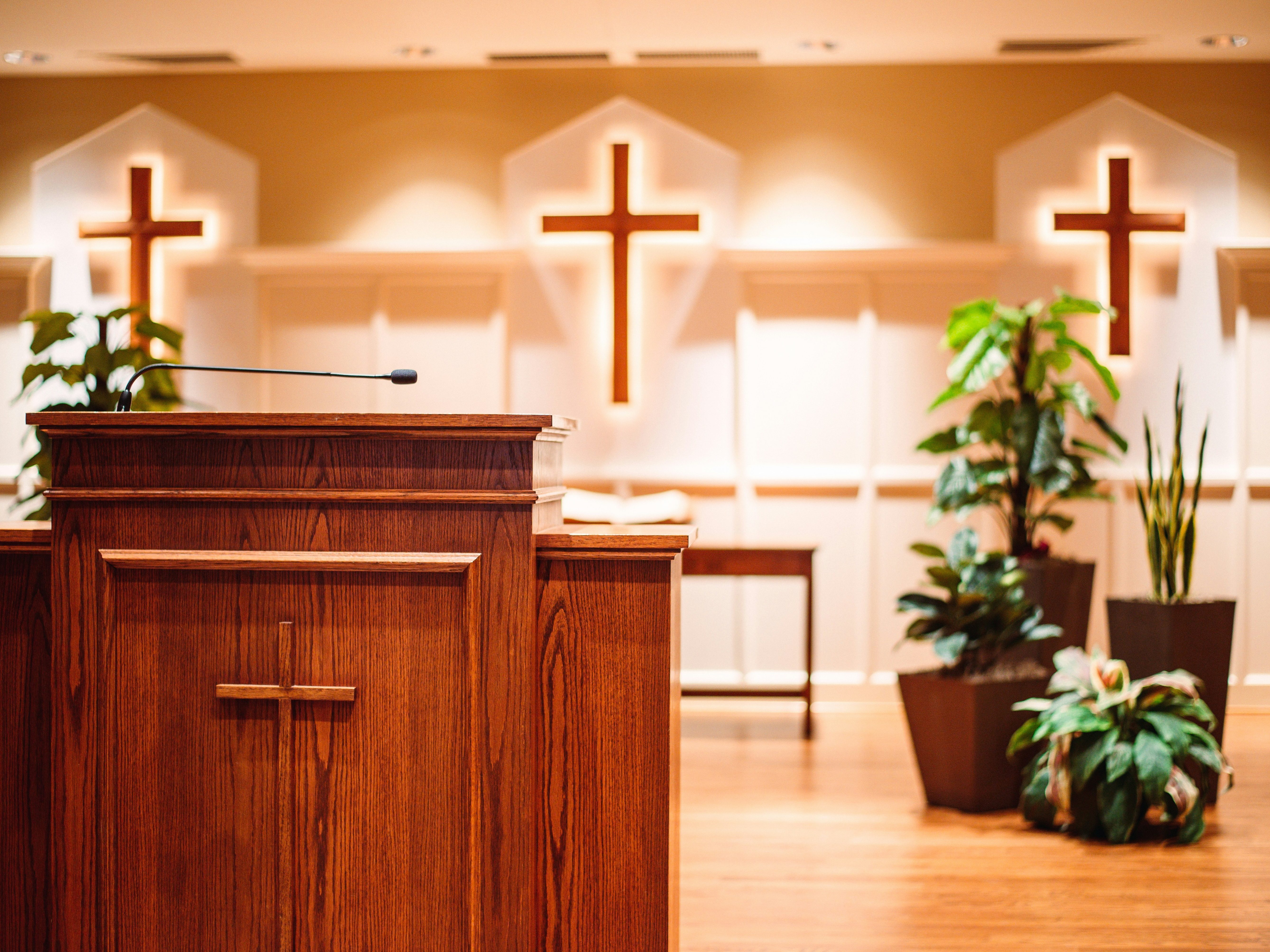 Wooden podium with a microphone in front of three illuminated crosses and decorative plants in a church interior.