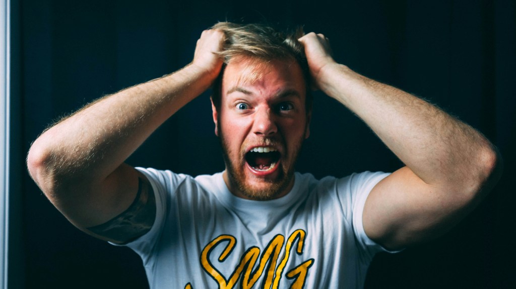 A man with short blond hair is screaming and pulling his hair in frustration, wearing a white t-shirt with yellow text, against a dark background.